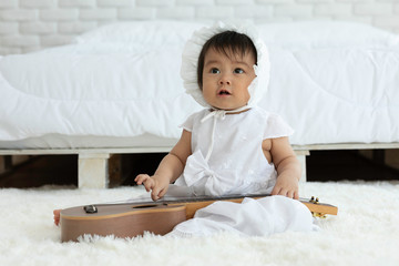 Cute Sweet Adorable Asian Baby wearing white dress sitting with guitar on Carpet smiling and playing with happiness emotional in cozy bedroom,Healthy Baby Concept