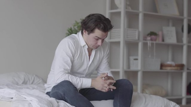 Sad young man alone at home or hotel room. The guy falling on his bed, he is sad, tired and exhausted. Mental injury, disorder, bad mood. Side view