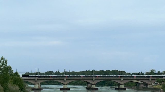 Static zoom in shot of a high-speed TGV train as it crosses a rail bridge over the river