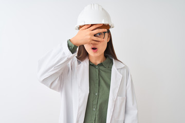 Young chinese engineer woman wearing coat helmet glasses over isolated white background peeking in shock covering face and eyes with hand, looking through fingers with embarrassed expression.