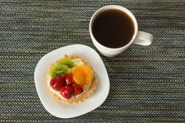 Cake with cream, raspberries, kiwi and orange and a cup of coffee on a black napkin. Pastry with fruits on top. Morning coffee. Copy space.