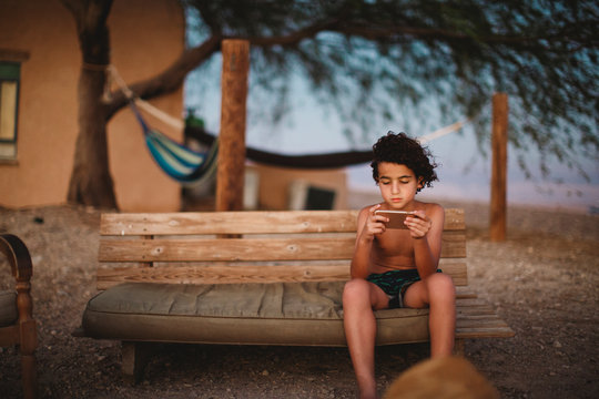 A young boy playing with his smartphone sitting on a bench in desert