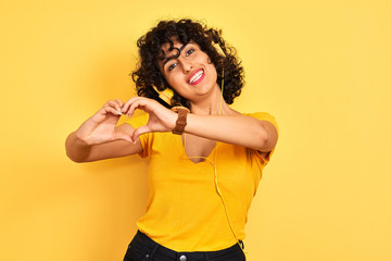 Fototapeta na wymiar Arab woman with curly hair listening to music using headphones over isolated yellow background smiling in love showing heart symbol and shape with hands. Romantic concept.