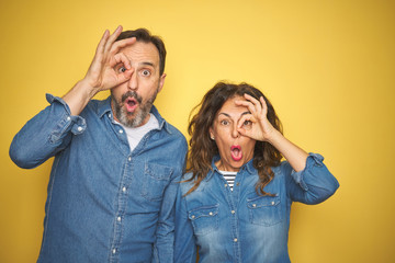 Beautiful middle age couple together standing over isolated yellow background doing ok gesture shocked with surprised face, eye looking through fingers. Unbelieving expression.