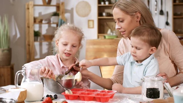Waist-up shot of young Caucasian mom with two kids sitting together at kitchen table with baking ingredients, condiments and utensils on it, and spooning batter into silicone cupcake mold
