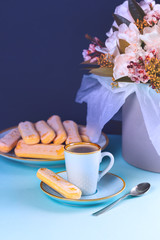 A cup of coffee on a blue saucer with Savoyardi cookies, against the backdrop of chic white tulips in a box.