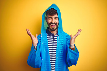 Young handsome man wearing rain coat with hood standing over isolated yellow background celebrating...