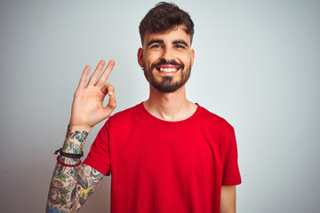 Fototapeta premium Young man with tattoo wearing red t-shirt standing over isolated white background smiling positive doing ok sign with hand and fingers. Successful expression.
