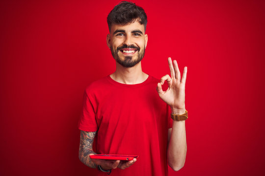 Young man with tattoo using tablet standing over isolated red background doing ok sign with fingers, excellent symbol