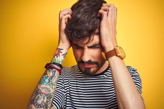 Young man with tattoo wearing striped t-shirt standing over isolated yellow background suffering from headache desperate and stressed because pain and migraine. Hands on head.