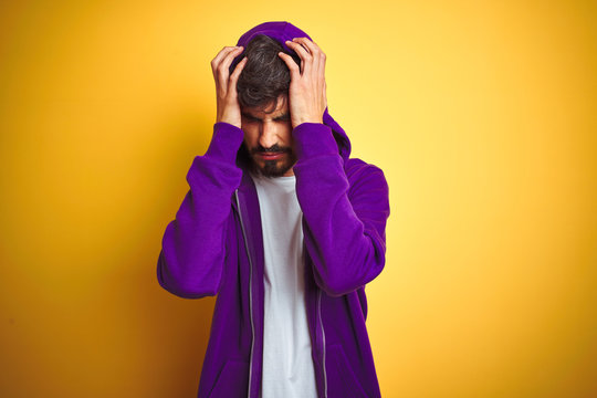 Young man with tattoo wearing purple sweatshirt with hood over isolated yellow background suffering from headache desperate and stressed because pain and migraine. Hands on head.