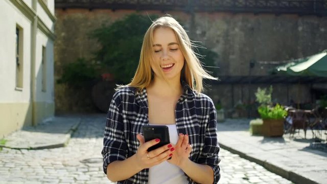 Enjoyable blonde young lady in trendy clothes using smartphone and touching her hair, walking on the street