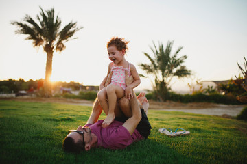Father laying on grass holding his laughing daughter up on his legs