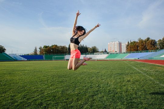 Attractive sporty happy brunette woman in pink shorts and top makes a high jump in sun rays at the stadium