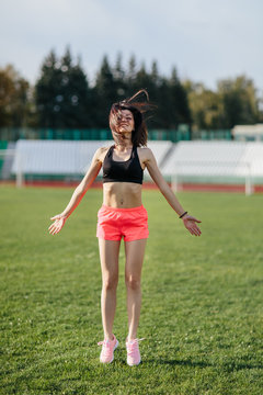 Attractive sporty brunette woman in pink shorts and top doing workout with jump rope in sun rays at the stadium