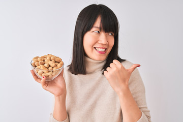 Young beautiful Chinese woman holding bowl with peanuts over isolated white background pointing and showing with thumb up to the side with happy face smiling