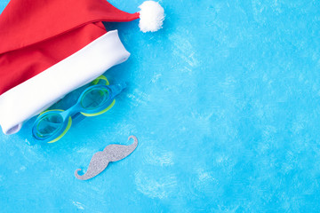 Hat of Santa Claus with goggles for swimming and a mustache. Christmas vacation, sandals and swimming glasses by water, slippers and pool goggles near swimming pool