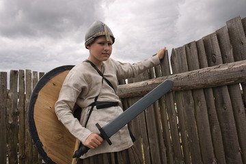 A teenager boy in a historical Viking costume with a helmet and a shield holding a sword standing in the middle of a wooden fortress at a historic festival.
