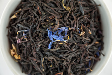 Dry black tea with blue flowers leaves in white bowl, top view, macro
