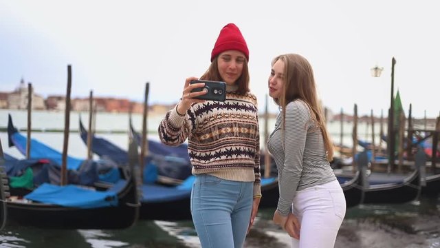 Two young women on the waterfront with boats are photographed on the phone and look at the photo