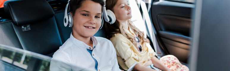 panoramic shot of boy listening music in headphones near sister in car