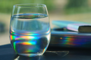 Transparent glass filled with water reflecting rainbow spots from a notebook in holographic cover