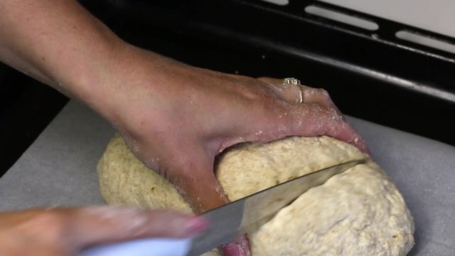 On the baking sheet is a lump of dough made from wholegrain flour. In the form of a loaf of bread. A woman makes incisions with a knife.