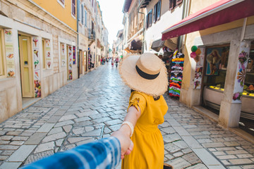 follow me concept. woman in yellow sundress in straw hat walking forward by small resort city street holding man hand