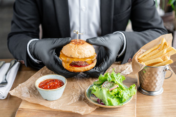 Young man in business suit and black gloves eating delicious juicy cheeseburger with beef chop....
