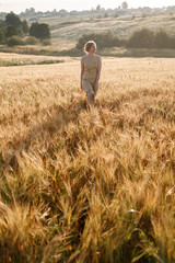 Naklejka na ściany i meble young pretty girl in yellow dress stands at field of ears in rays of rising sun. grove and village in background
