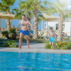 Smiling Caucasian man and his son are jumping in to the swimming pool at resort on family vacation.