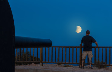 A man looks at the full moon with partial lunar eclipse. A gun is aimed at the horizon. The place is a Spanish defensive facility near the city of Cartagena.