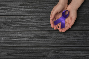 Woman holding purple awareness ribbon on black wooden background, top view with space for text