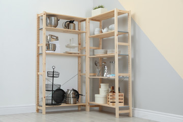 Wooden shelving units with kitchenware near color wall. Stylish room interior