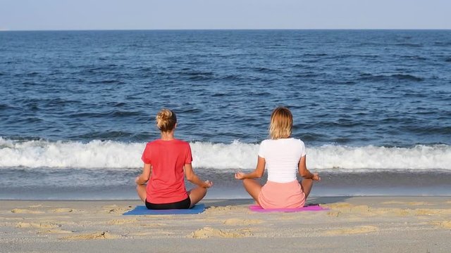 Young women practicing yoga on the beach at sunset. Girls meditating, sitting in lotus pose on the sea shore.