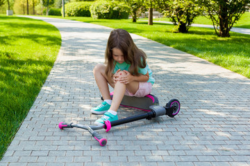 Cute little girl sitting on the ground after falling off her scooter at summer park. Child getting hurt while riding a kick scooter. Active healthy outdoor sport for young child. Fun activity for kid