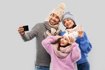 family, christmas and technology concept - happy mother, father and little daughter in winter clothes taking selfie by smartphone over grey background