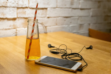 A bottle with juice and a straw is on the table. Cell phone with headphones lying on the table. Youth set, lifestyle