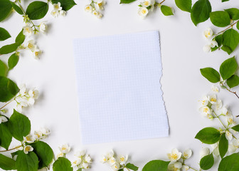 Blank piece of paper in frame of white flowers and green leaves on white background, top view. Spring card with copy space for text.