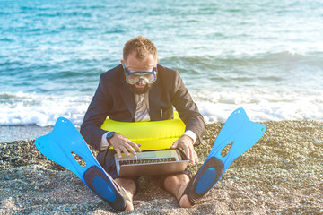 Funny businessman wearing snorkeling tools is working on the beach with laptop