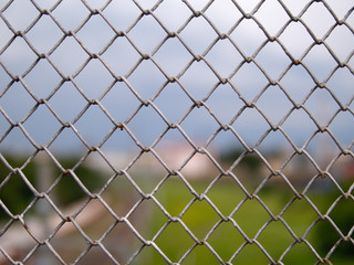 Fototapeta premium Side view and selective focus of grey wire mesh fence with blurred background of cloudy sky, green grass, buildings