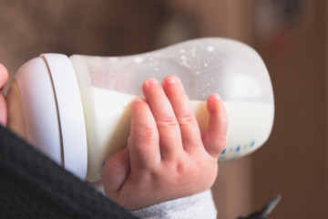 A small child drinks milk from a plastic bottle, holding with hands. Food for the child. Side view. 
