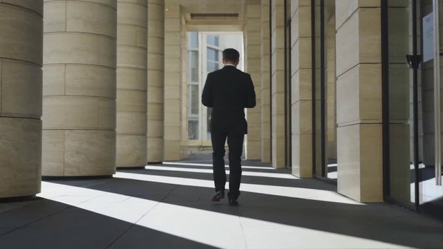 Full length follow shot back view of unrecognizable middle aged businessman in black suit walking down street in slow motion and using cell phone