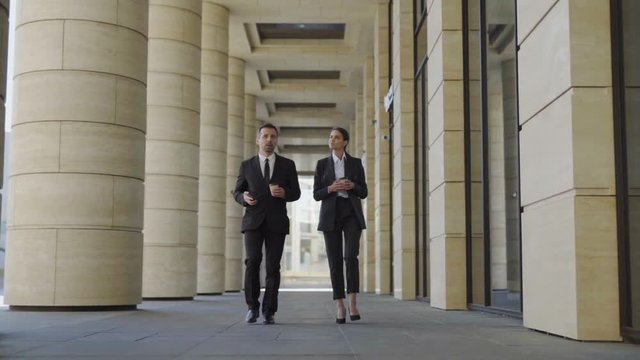 Full length slow motion shot of two businesspeople in formal suits, middle aged man and young woman walking down street with takeaway coffee cups, looking around and discussing architecture