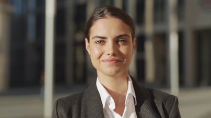 Head and shoulders slow motion portrait of beautiful and confident young woman in formal suit standing outdoors and posing for camera with happy smile