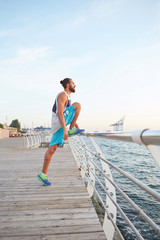 Portrait of young bearded sporty guy doing morning exercises by the sea, stretching for legs, warm-up after run, leads healthy active lifestyle. Fitness male model.