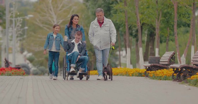 Parents walk the path with the children and the dog. Son in a wheelchair rides with his family along the avenue.