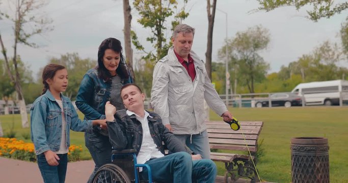 Sister and mother are taking brother in a wheelchair. Happy family with disabled teenager together walking.