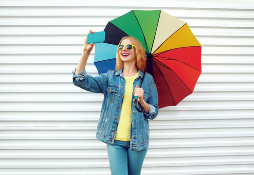 Happy smiling woman taking selfie picture by phone with colorful umbrella in city on white wall background