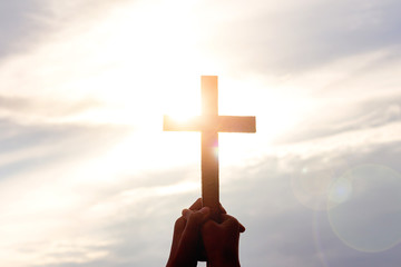 human hands praying to the GOD while holding a crucifix symbol with bright sunbeam on the sky